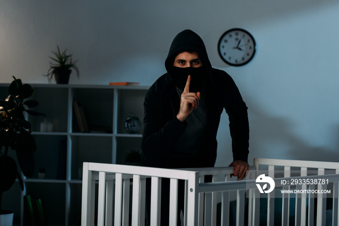 Kidnapper in mask and black hoodie showing hush sign beside crib