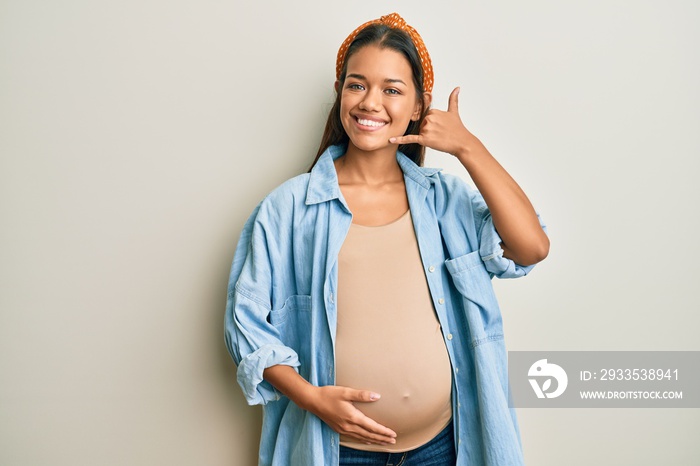 Beautiful hispanic woman expecting a baby, touching pregnant belly smiling doing phone gesture with hand and fingers like talking on the telephone. communicating concepts.