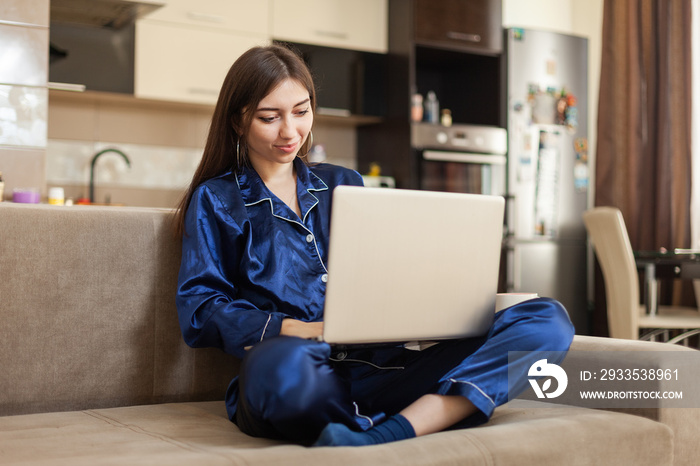 Young cheerful woman in nightie looks into the laptop screen while sitting on the sofa in the living room