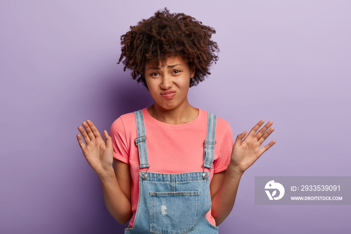 Thank yoy but no. Displeased dark skinned woman receivs disturbing suggestion, waves palms in rejection, grimaces unpleasantly, wears casual pink t shirt and dungarees, poses in studio alone