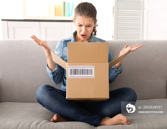 Beautiful young woman opening box with parcel while sitting on sofa at home