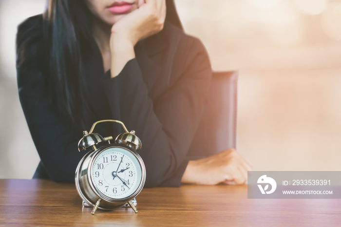 wasting time concept with Asian business woman feeling tired and bored waiting for someone coming late at work and looking at alarm clock on desk