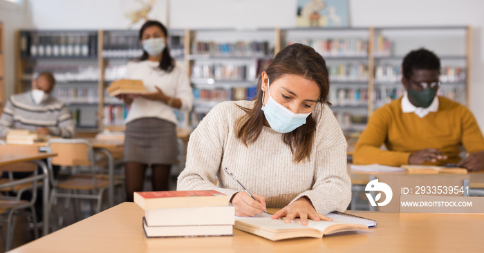Young latin american woman wearing protective face mask studying in university library, making notes in workbook. New normal during pandemic..