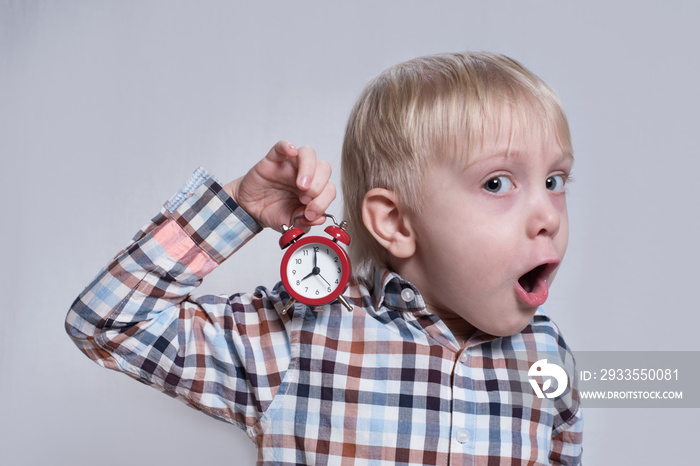 Little blond boy with a red alarm clock in his hands. Surprised face. Morning concept. Light background