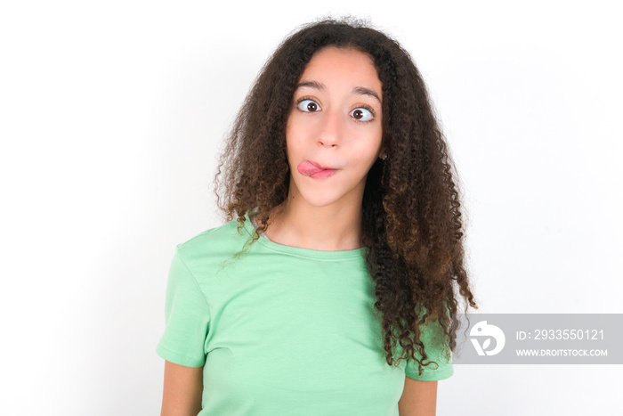 Funny Teenager girl with afro hairstyle wearing green T-shirt over white wall makes grimace and crosses eyes plays fool has fun alone sticks out tongue.