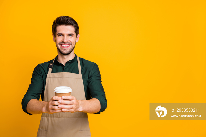 Photo of cheerful positive handsome attractive man holding paper cup with hands recommending you to taste this sort of coffee wearking apron isolated over yellow vivid color background