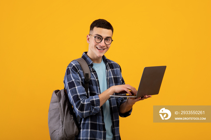 Intelligent young student with backpack using laptop pc on orange studio background