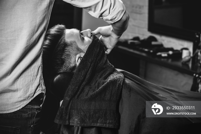 Client during beard shaving in barbershop. Young handsome barber and attractive bearded man. Black and white or colorless photo. Hairstyle, salon, hairdresser, barber shop, lifestyle concept