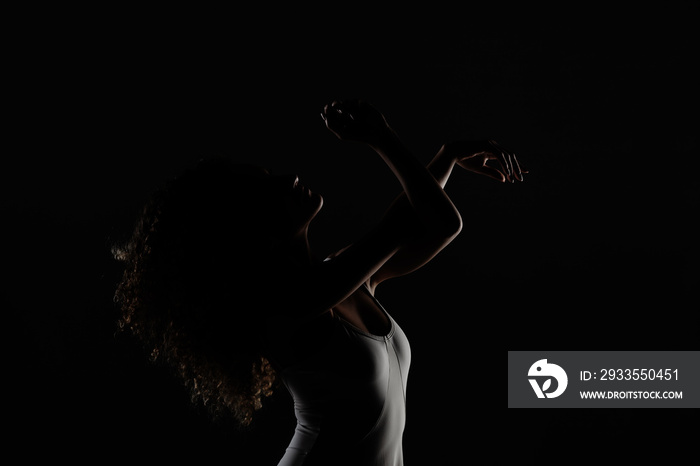 Girl with curly hair making ballet poses. Side lit silhouette of ballerina in white dress against black background.