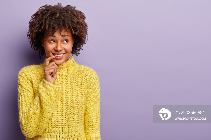 Optimistic glad woman keeps hand near broad smile, shows white teeth, focused aside, dreams about new desirable thing, wears yellow knitted jumper, models over purple background. Emotions concept