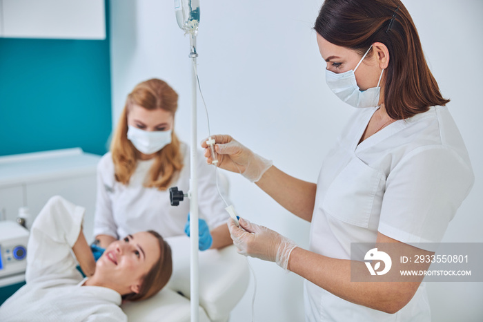 Brunette physician in white uniform and gloves looking to the intravenous vitamin drip in spa salon