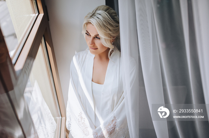 Beautiful blonde bride stands near the window in a white coat and lingerie. The morning of the bride. Wedding portrait of a cute girl in a hotel. Concept of quarantine, self-isolation. Stay home.