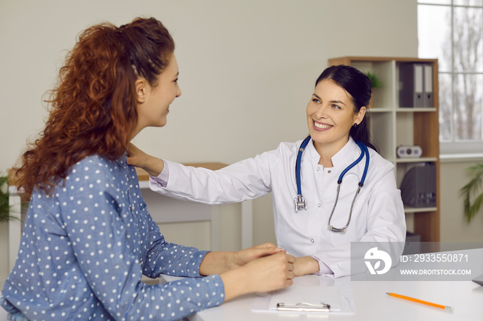 Happy supportive doctor talking to patient. Cheerful friendly doctor in white coat with stethoscope sitting at desk, smiling and touching woman’s shoulder. Health checkup, medical consultation concept