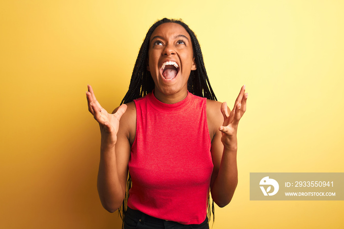 African american woman wearing red casual t-shirt standing over isolated yellow background crazy and mad shouting and yelling with aggressive expression and arms raised. Frustration concept.