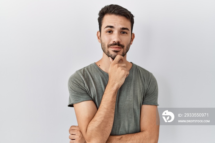 Young hispanic man with beard wearing casual t shirt over white background looking confident at the camera smiling with crossed arms and hand raised on chin. thinking positive.