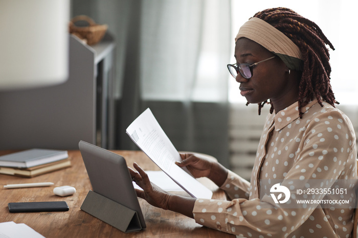 Side view portrait of young African-American woman wearing glasses and using digital tablet on stand while working from home, copy space