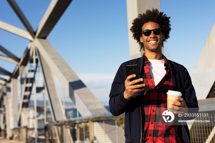 Smiling black man holding coffee to go. Happy African man using the phone outside.