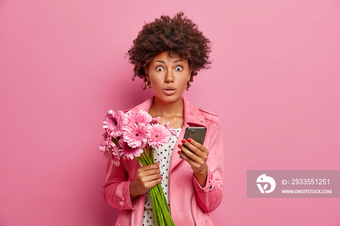 Photo of surprised female model holds bouquet of flowers modern smartphone receives unexpected congratulation on birthday dressed in stylish clothes. Women spring time special occasion concept