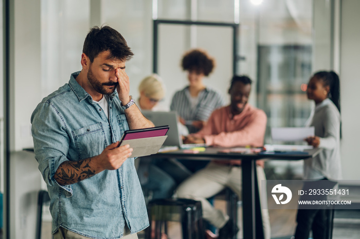 Stressed business man using tablet in the office with his team working in the back