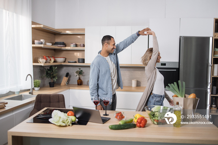 Young attractive couple of man and woman cooking breakfast together in morning at kitchen, happy family, romantic, healthy food, freelancer at work on laptop, dancing to music, having fun, laughing.