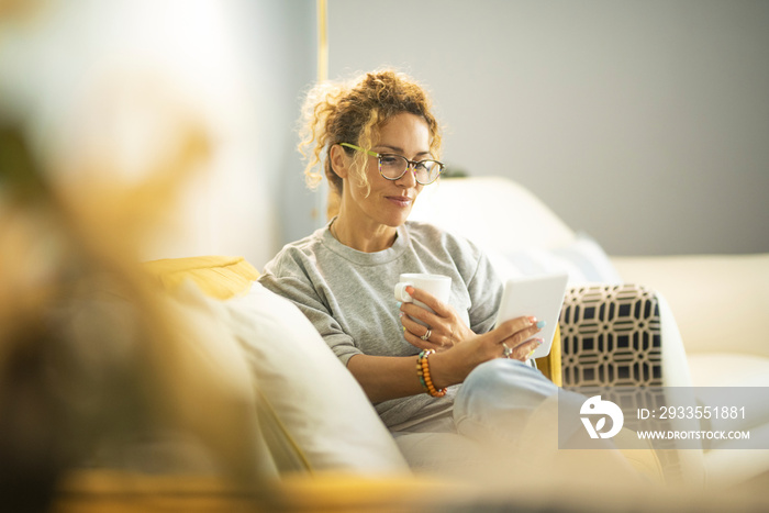 Smiling middle-aged Caucasian woman sit on couch in living room browsing wireless Internet on tablet, happy modern pretty female relax on sofa at home using pad device, yellow, technology concept