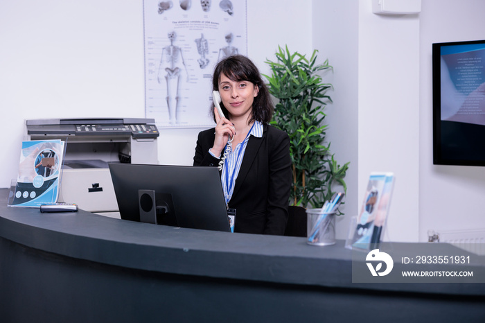 Portrait of smiling hospital receptionist answering call from patient to make an appointment for clinical consult at private clinic. Professional looking woman working at front desk using telephone.