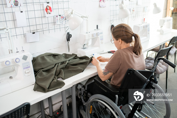 Young disable female in casualwear sitting in wheelchair by workplace inside contemporary workshop