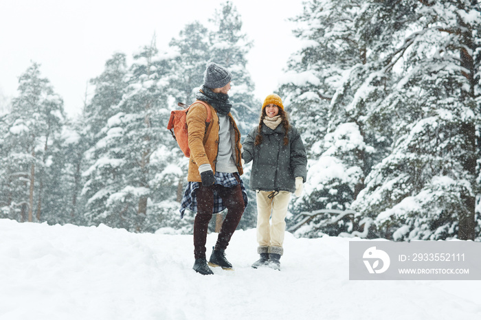 Happy young couple of hikers holding hands and looking at each other while walking under snow in winter forest