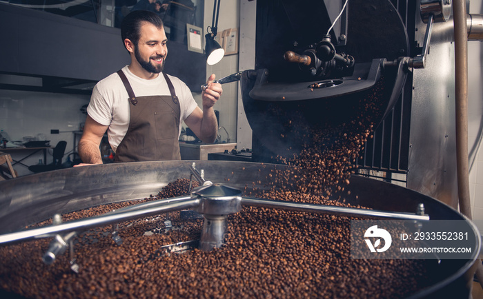 Portrait of cheerful bearded master having job at factory. He controlling brown beans pouring into cooler machine from large coffee roaster. Work concept