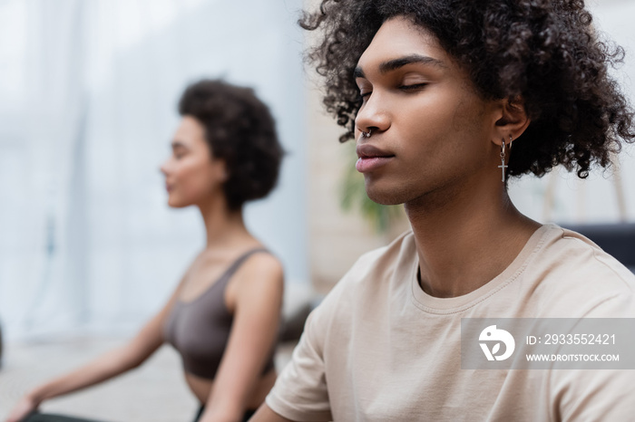 Young african american man meditating near blurred girlfriend at home.