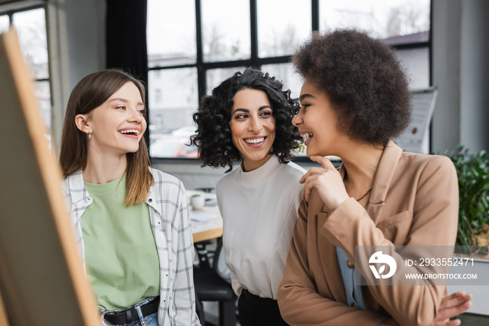 Cheerful interracial businesswoman talking near blurred board in office.