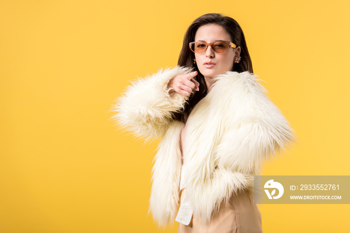 girl in faux fur jacket and sunglasses pointing with finger at camera isolated on yellow