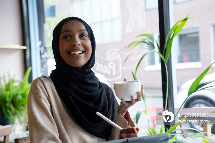 Smiling young woman in hijab drinking coffee in cafe