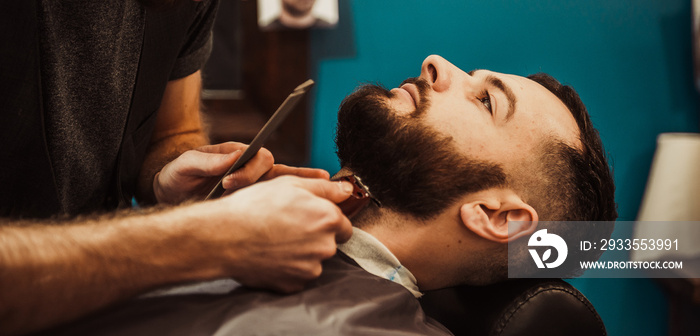 A professional barber cuts his beard to a young hipster man.