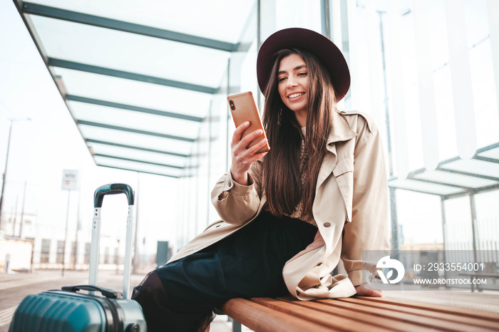 Beautiful positive girl traveler in coat and hat on his head sitting at a bus stop waiting for a bus and uses a smartphone. Mobile technology concept