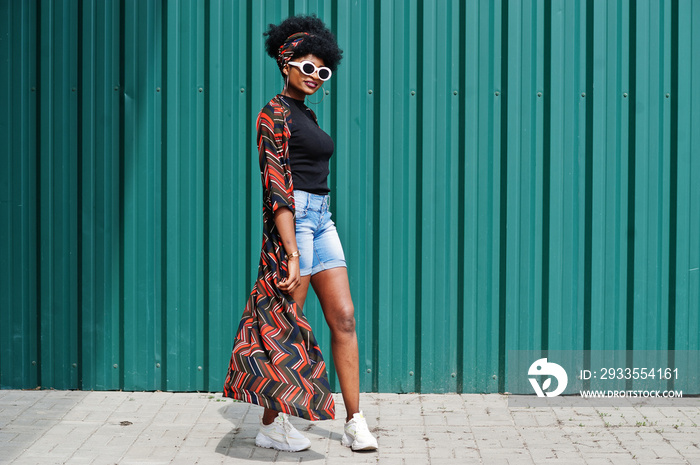 African woman with afro hair, in jeans shorts and white sunglasses posed against green steel wall.