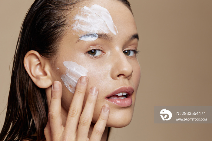 young woman applying a soothing face mask cosmetic beige background