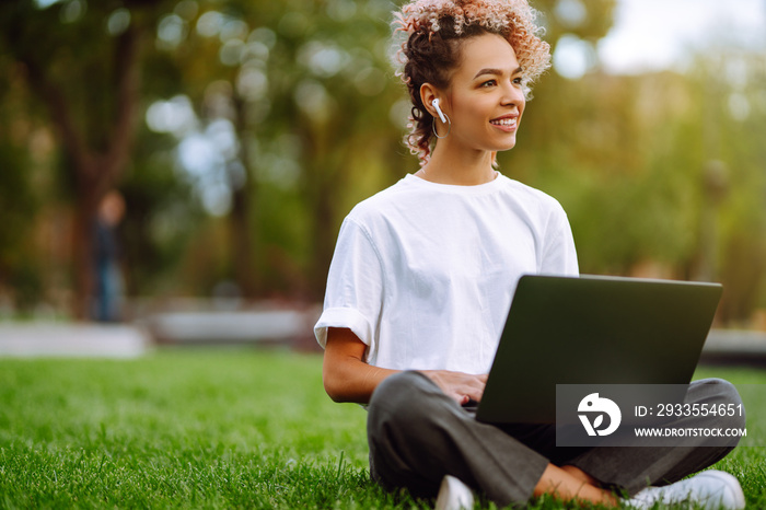 Young woman sitting on the grass  in a city park and working on a laptop. Freelance business concept. Technology and modern lifestyle concept.