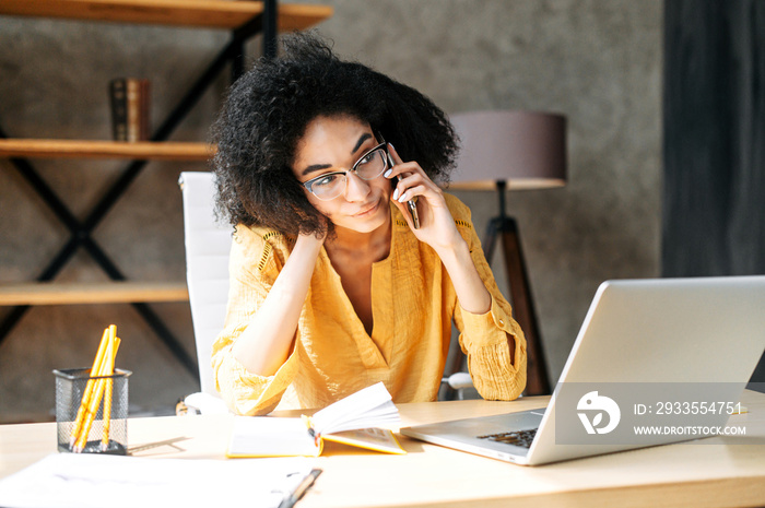 A tired african american woman is talking sitting at modern office. Unhappy young employee takes calls at work