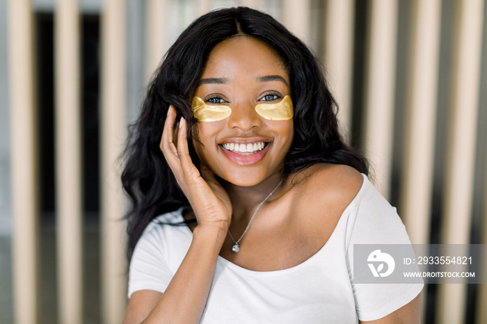 Beautiful young African woman applies gold eye mask, medical eye antiwrinkle patches. Portrait of beauty dark skinned model with natural nude make up cares about her skin. Spa, skincare at home