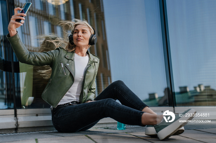 Beautiful young blonde woman in headphones wearing casual clothes sitting on the ground enjoying music, makes selfie on weekend. Cheerful italian hipster girl relaxing outdoors. Youth, leisure.