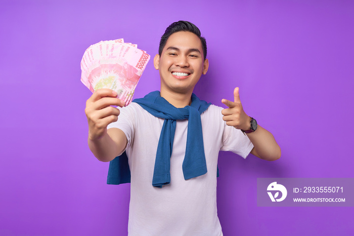 Smiling young Asian man wearing white t-shirt pointing fingers at cash money in rupiah banknotes isolated over purple background. people lifestyle concept