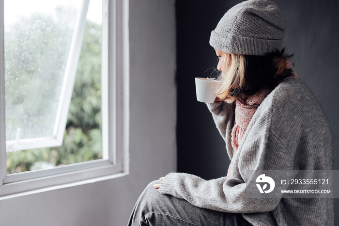 Lonely girl looking outside through the window while enjoying a cup of coffee