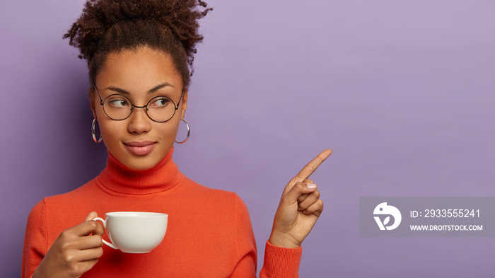 Serious female office worker has coffee break, holds white mug of hot beverage, points aside on blank space, has self confident face expresson, wears glasses and turtleneck, isolated over violet wall