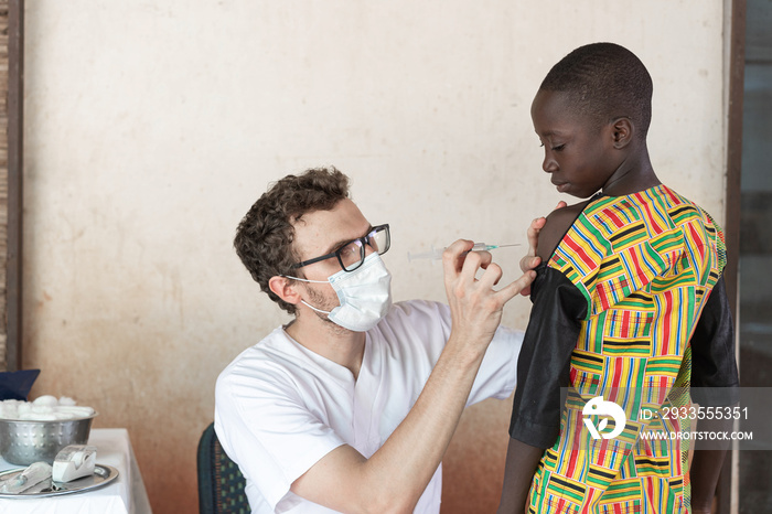 Medical professional with protective face mask and glasses preparing the injection site on the arm of a brave black African schoolboy in oder to to administer a first dose of antimalarial vaccine