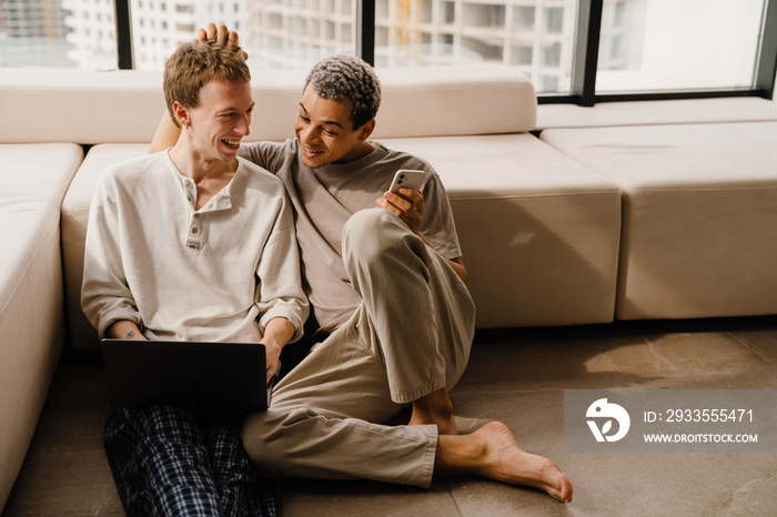 Happy gay couple using cellphone and laptop while resting at home