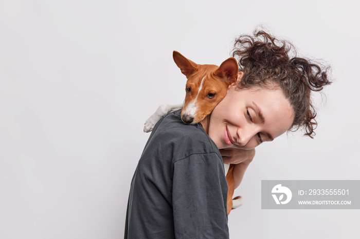 Sideways shot of adult girl carries puppy which embraces her around neck walkk together isolated over white background. Female dog lover poses with pet looks away likes animals. Care and love