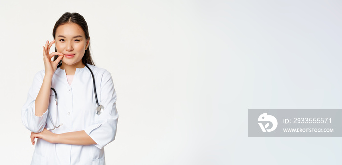 Taboo, medical confidentiality concept. Young asian doctor, female physician showing mouth silence, taboo or zip gesture, promise not tell, standing over white background