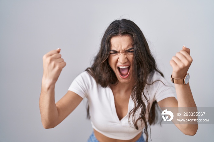 Young teenager girl standing over white background angry and mad raising fists frustrated and furious while shouting with anger. rage and aggressive concept.