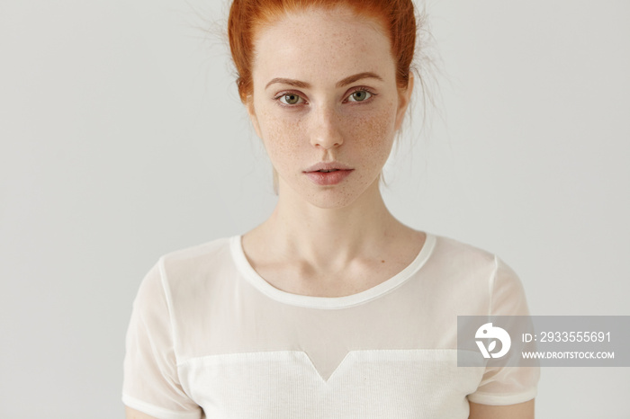 Cropped studio shot of beautiful Caucasian female with red hair and freckles wearing white top, standing isolated at blank wall, having serious look, staring at camera. People and lifestyle concept